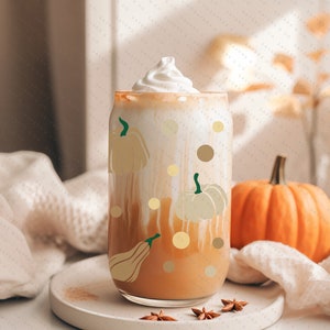 May include: A glass of pumpkin spice latte with whipped cream and a pumpkin spice design. The glass is decorated with gold pumpkins and dots. A small orange pumpkin sits on a white surface next to the glass.