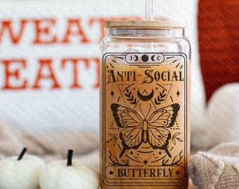May include: A clear glass tumbler with a bamboo lid and a clear straw. The tumbler features a decorative label with the words "ANTI-SOCIAL BUTTERFLY" and a butterfly illustration. The tumbler is filled with a light brown liquid.