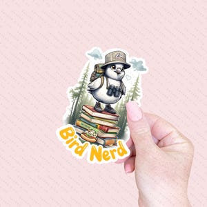 May include: A white bird wearing a hat and binoculars stands on a stack of books. The bird is holding a hot dog in its beak. The text "Bird Nerd" is written in yellow letters below the bird.