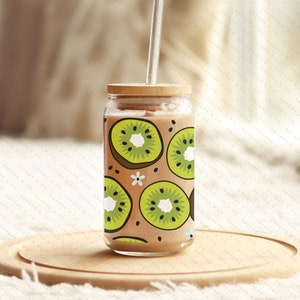 May include: A glass jar with a brown wooden lid and a clear plastic straw. The jar is filled with a brown liquid and has a pattern of green kiwi slices on the outside.