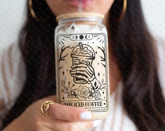 May include: A glass jar with a black and white illustration of a hand holding a cup of iced coffee. The illustration is surrounded by flowers and stars. The text "THE ICED COFFEE" is printed below the illustration.