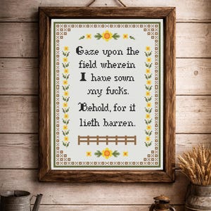 May include: A framed cross-stitch artwork with a rustic aesthetic. The text reads "Gaze upon the field wherein I have sown my fucks. Behold, for it lieth barren." The frame is brown, and the background is a light cream color.