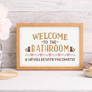 Könnte beinhalten: Ein gerahmtes Kreuzstichschild mit den Worten "WELCOME TO THE BATHROOM" und "A CAT WILL BE WITH YOU SHORTLY". Das Schild hat einen Holzrahmen und zeigt Katzen- und Herzmotive. Eine Vase mit rosa Blumen und ein Stück Seife sind ebenfalls zu sehen.