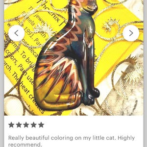 May include: A colorful glass cat figurine with a seated pose. The cat features a blend of yellow, orange, blue, and brown hues. The background includes text and a yellow and gold design.