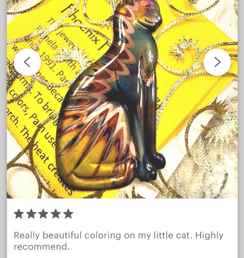 May include: A colorful glass cat figurine with a seated pose. The cat features a blend of yellow, orange, blue, and brown hues. The background includes text and a yellow and gold design.