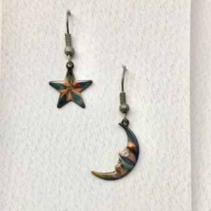 May include: A pair of earrings featuring a crescent moon and a star. The moon and star are both made of metal and have a multi-colored enamel design. The earrings are silver in color.
