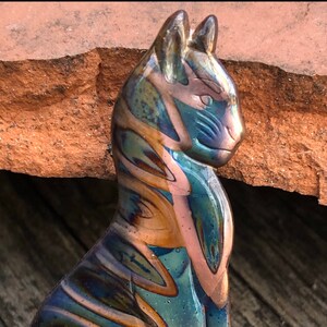 May include: A glass cat figurine with a glossy finish. The cat is primarily blue and gold, with streaks of pink and green. The figurine is set against a backdrop of a rough, reddish-brown surface.