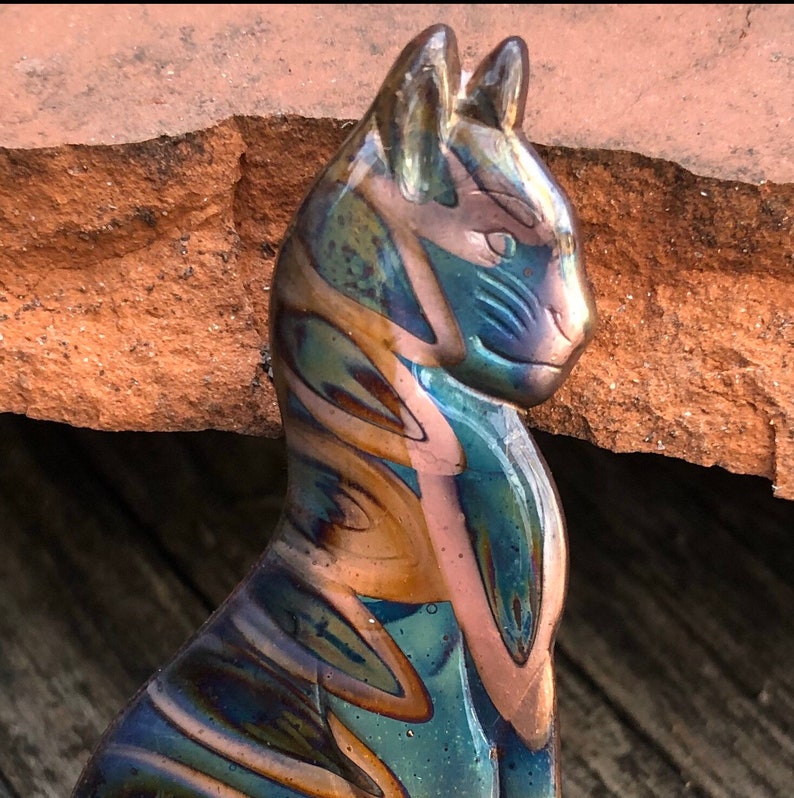 May include: A glass cat figurine with a glossy finish. The cat is primarily blue and gold, with streaks of pink and green. The figurine is set against a backdrop of a rough, reddish-brown surface.