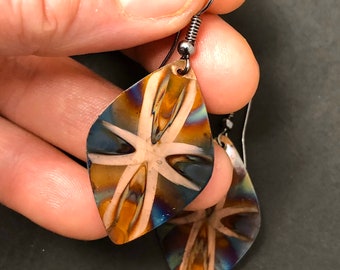 Puede incluir: Un par de pendientes en forma de lágrima con un diseño abstracto único. Los pendientes presentan una mezcla de colores, incluyendo azul, dorado y blanco, con un patrón en forma de estrella. Los ganchos para las orejas son de metal oscuro.