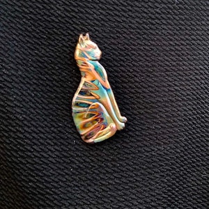 May include: A sitting cat-shaped pendant with iridescent colors of blue, gold, and purple. The cat is detailed with a smooth, polished finish. The pendant is likely made of glass or a similar material.