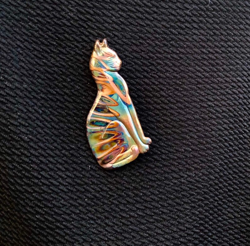May include: A sitting cat-shaped pendant with iridescent colors of blue, gold, and purple. The cat is detailed with a smooth, polished finish. The pendant is likely made of glass or a similar material.