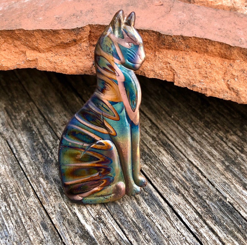 May include: A colorful, iridescent glass cat figurine in a seated position. The cat features shades of blue, green, and bronze, with swirling patterns. The figurine is displayed on a weathered wooden surface.