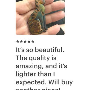 May include: A handcrafted cat figurine in shades of green, brown, and gold, held in a hand. The cat is in a seated position with intricate detailing. The text on the image says, "It's so beautiful. The quality is amazing, and it's lighter than I expected. Will buy another piece!"