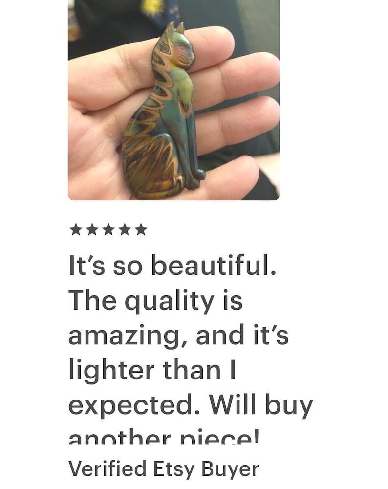 May include: A handcrafted cat figurine in shades of green, brown, and gold, held in a hand. The cat is in a seated position with intricate detailing. The text on the image says, "It's so beautiful. The quality is amazing, and it's lighter than I expected. Will buy another piece!"