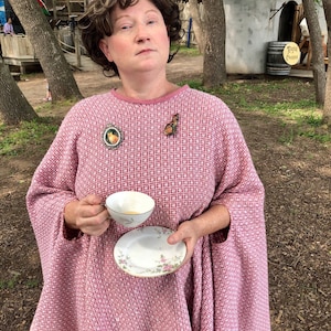 May include: A woman wearing a pink woven cape with a matching pink dress. She is holding a teacup and saucer. The cape has a geometric pattern. She is wearing a brown wig and a pink hat. There are two brooches on the cape.