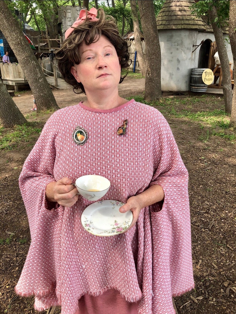 May include: A woman wearing a pink woven cape with a matching pink dress. She is holding a teacup and saucer. The cape has a geometric pattern. She is wearing a brown wig and a pink hat. There are two brooches on the cape.