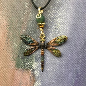 May include: A gold dragonfly pendant necklace with a green stone bead and gold accents. The dragonfly has iridescent wings with shades of blue, green, and gold.