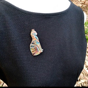 May include: A colorful cat-shaped brooch with a unique, swirled design in shades of blue, orange, and gold. The brooch is made of a shiny material and is attached to a black dress. The cat is sitting upright, with its tail curled.
