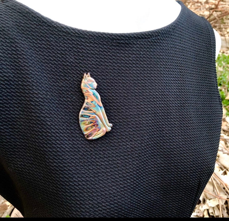 May include: A colorful cat-shaped brooch with a unique, swirled design in shades of blue, orange, and gold. The brooch is made of a shiny material and is attached to a black dress. The cat is sitting upright, with its tail curled.
