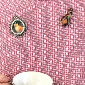 May include: Two cat-themed brooches on a red and white patterned fabric. One brooch is oval with a cat portrait and pearl-like trim. The other is a small, detailed cat figurine in shades of brown and orange.