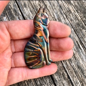 May include: A glass cat figurine with a seated pose. The cat features a blend of blue, green, and bronze colors with a swirled pattern. The figurine is held in a hand, showcasing its size and detail.