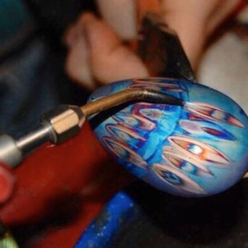 May include: A close-up of a glass art piece in the process of being crafted. The piece is a vibrant blue with intricate patterns in shades of orange and white. A torch is being used to shape the glass, highlighting the artistry involved.