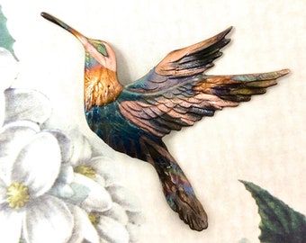 May include: A detailed, metal hummingbird sculpture with iridescent colors of blue, bronze, and pink. The bird is in mid-flight, wings outstretched, and has a long, slender beak. The background features a floral design.