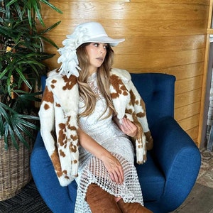 May include: A woman wearing a white dress with a lace overlay, a brown cow print jacket, and brown over-the-knee boots. She is sitting on a blue armchair and wearing a white hat with a large flower.