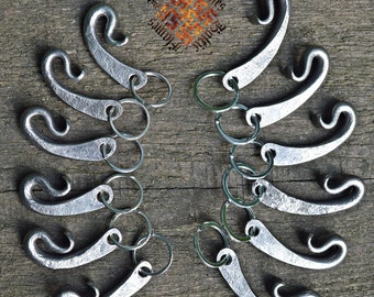 Può includere: Portachiavi in metallo forgiato a mano con un design a gancio curvo. Ogni portachiavi è attaccato a un anello di metallo. I portachiavi sono disposti su una superficie di legno invecchiato. Il testo "Baltic Flames" è al centro.