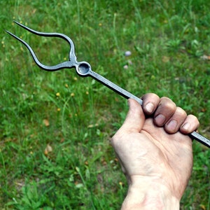 May include: Hand-forged metal grilling fork with a two-pronged end and a ring on the handle. The dark gray fork is held against a backdrop of green grass.