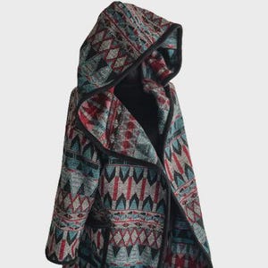 May include: A hooded jacket with a geometric pattern in shades of teal, red, and gray. The jacket has a black trim and a draped, open front design. The hood is prominent, and the overall style is bohemian.