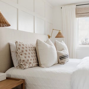 May include: A bedroom scene featuring a bed with white bedding and decorative pillows. The pillows have various patterns and textures, including floral and striped designs. Woven lamps hang above the bed, and a window with white curtains is visible.