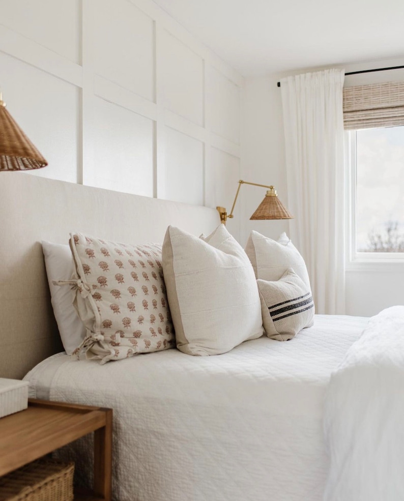 May include: A bedroom scene featuring a bed with white bedding and decorative pillows. The pillows have various patterns and textures, including floral and striped designs. Woven lamps hang above the bed, and a window with white curtains is visible.