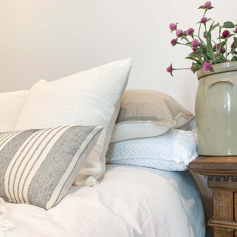 May include: A white bed with a white duvet cover and several pillows. The pillows are arranged in a stack, with a striped pillow on top. A vase with pink flowers sits on a wooden nightstand next to the bed.