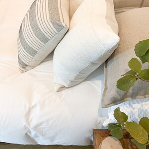 May include: A white bed with a white duvet cover and three pillows. The pillows are a striped black and white pillow, a white pillow with a textured surface, and a beige pillow. A wooden end table with a round wooden coaster sits next to the bed.
