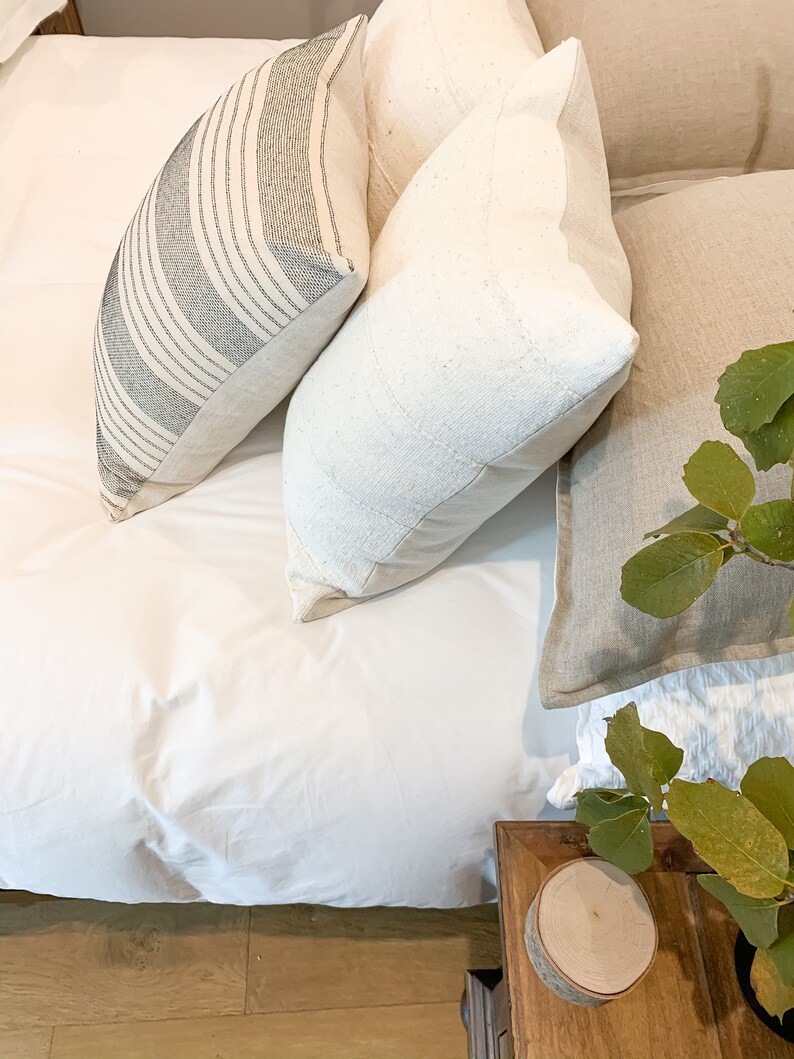 May include: A white bed with a white duvet cover and three pillows. The pillows are a striped black and white pillow, a white pillow with a textured surface, and a beige pillow. A wooden end table with a round wooden coaster sits next to the bed.