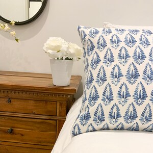 K&ouml;nnte beinhalten: Ein wei&szlig;es Bett mit einem blau-wei&szlig; gemusterten Kissen. Eine wei&szlig;e Keramikvase mit wei&szlig;en Blumen steht auf einem h&ouml;lzernen Nachttisch.