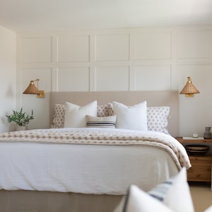 May include: A bedroom scene featuring a bed with a beige headboard, white bedding, and decorative pillows. A patterned throw blanket adds a touch of color. Woven sconces flank the headboard, and a wooden nightstand is visible.