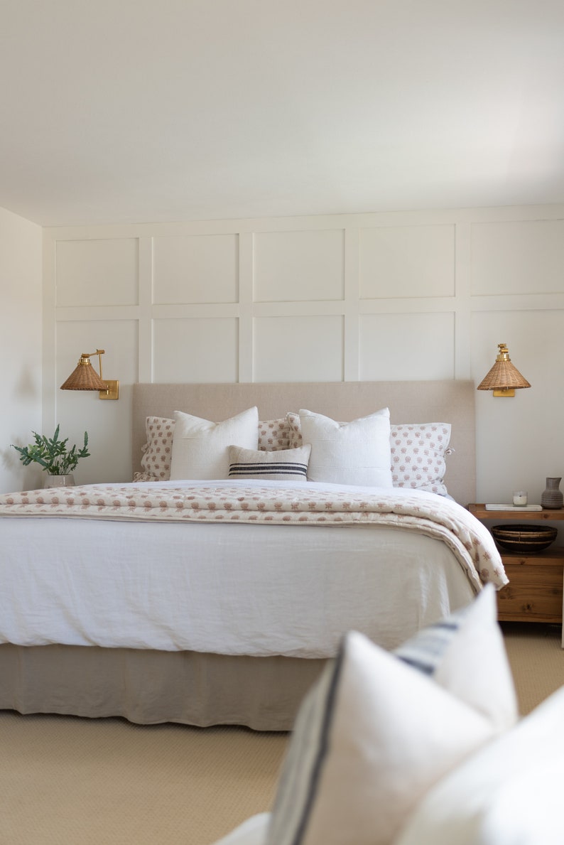 May include: A bedroom scene featuring a bed with a beige headboard, white bedding, and decorative pillows. A patterned throw blanket adds a touch of color. Woven sconces flank the headboard, and a wooden nightstand is visible.