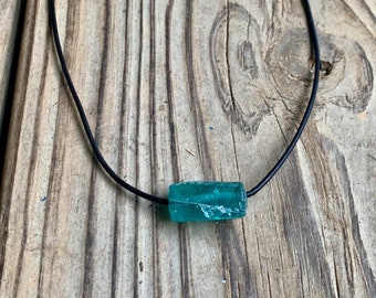 May include: A turquoise blue glass bead necklace on a black leather cord. The bead is rectangular and has a rough, textured surface.