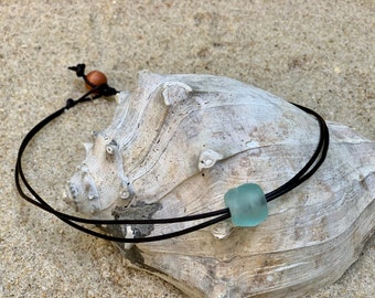 May include: A simple necklace with a light blue sea glass bead and a brown wooden bead. The necklace is made of black leather cord and is displayed on a seashell.