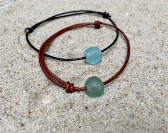 May include: Two leather bracelets with sea glass beads. One bracelet is brown leather with a green sea glass bead. The other bracelet is black leather with a light blue sea glass bead.