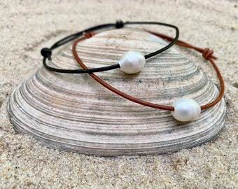 May include: Two leather cord necklaces with white pearl accents. The necklaces are displayed on a seashell against a sandy background.