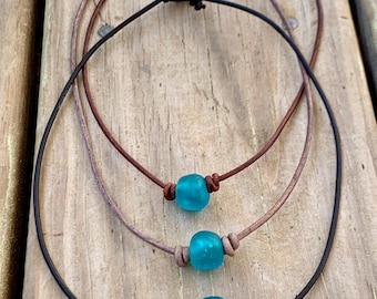 May include: Three leather necklaces with turquoise blue glass beads. The necklaces are layered on a wooden surface.