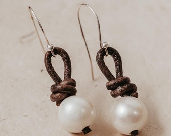 May include: A pair of brown leather earrings with a knotted design and a white pearl hanging from each earring.