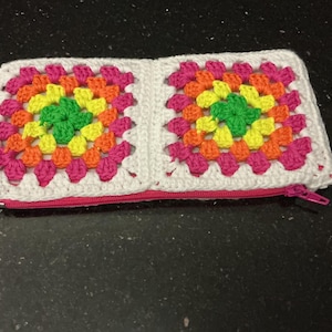 May include: A rectangular crochet pouch featuring two granny square motifs in vibrant shades of pink, orange, yellow, and green, bordered by white yarn. The accessory includes a bright pink zip closure along the bottom edge of the design.