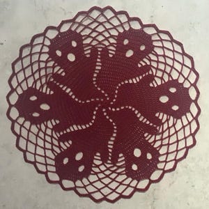 Op de afbeelding: Bordeaux gehaakte doily met een cirkelvormig ontwerp. Het midden heeft een gestileerd bloemmotief met opengewerkte details. De buitenrand heeft een geschulpte rand, wat een decoratief tintje geeft. Dit handgemaakte item is geschikt voor woondecoratie.