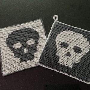 Könnte beinhalten: Zwei quadratische Häkel-Topflappen mit Totenkopfmotiv. Ein Topflappen ist hellgrau mit einem dunkelgrauen Totenkopf, der andere ist dunkelgrau mit einem hellgrauen Totenkopf. Jedes Stück hat eine kleine Häkelschlaufe an der oberen Ecke zum Aufhängen.