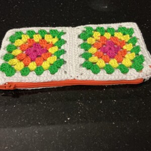 May include: A rectangular crochet pouch featuring two granny square motifs in vibrant shades of pink, orange, yellow, and green, bordered by white yarn. The pouch is secured with a bright orange zip closure along the bottom edge.