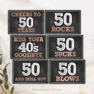 May include: Six black chalkboard signs with white chalk writing and rose gold accents. The signs feature the text "Cheers to 50 Years", "50 Rocks", "Kiss Your 40's Goodbye", "50 Sucks", "50 And Still Hot", and "50 Blows".